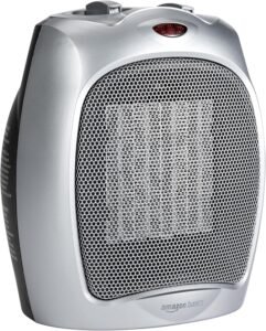Silver ceramic heater with a large front metal grille, visible ceramic heating element behind the mesh, and a small red indicator light near the top; “amazon basics” logo on the lower front.